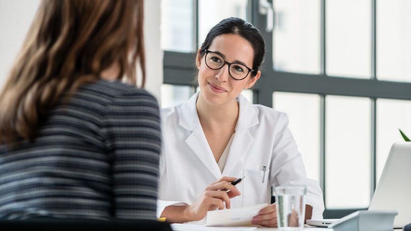 Eine Frau mit zusammengebundenen Haaren und Brille hält eine Kugelschreiber in der rechten Hand und ein Blatt Papier in der linken und berät eine andere Frau.
