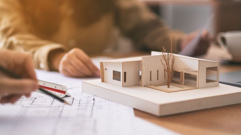 Mehrere Personen am Tisch mit einem physischen Architekturmodell, Bauzeichnungen und Papieren, die gemeinsam das Modell und Pläne im Büro betrachten und besprechen.