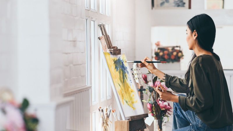 Künstlerin arbeitet in einem Atelier an einem abstrakten Acrylgemälde auf einer Staffelei. Im Hintergrund sind weitere Leinwände und Malutensilien zu sehen.