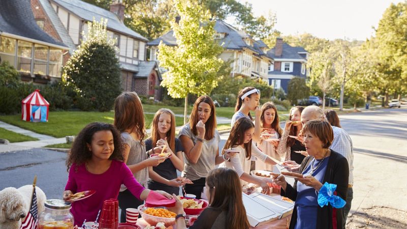 Menschen bedienen sich an einem Buffet bei einem Nachbarschaftsfest im Freien. Sie stehen gemeinsam an langen Tischen und kommen in ungezwungener Atmosphäre ins Gespräch.
