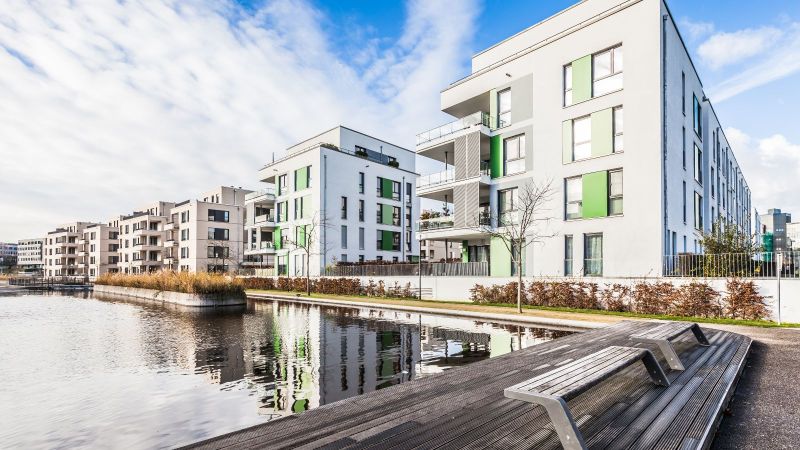 Moderne Neubausiedlung mit weißen Wohngebäuden und Balkonen an einem Kanal. Grünflächen und die klare, geometrische Architektur prägen das Wohnviertel.