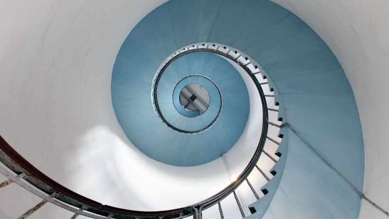 Blick von unten an einer spiralförmig verlaufenden Treppe entlang, die hoch in einen Turm führt.
