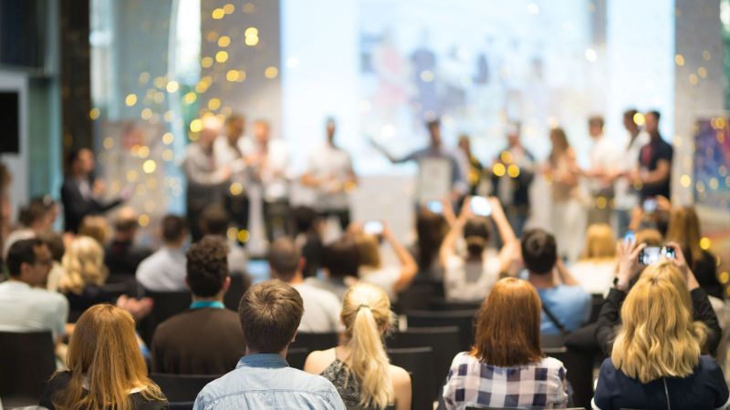 Ein junges Business-Team erhält bei einem Wettbewerb für das beste Geschäftsprojekt eine Auszeichnung. Die Veranstaltung steht unter dem Motto Wirtschaft, Unternehmertum und Preisverleihung. Der Fokus liegt auf den nicht erkennbaren Personen im Publikum.