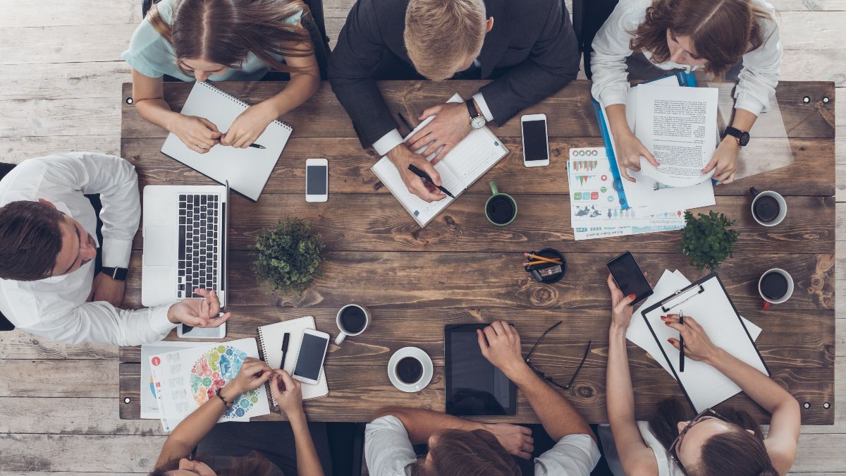Draufsicht auf einen Tisch mit Unterlagen, Laptops, Tablets, Smartphones und Kaffeetassen. Um den Tisch sitzen sieben Personen und diskutieren. | &copy;Pandora Studio - stock.adobe.com