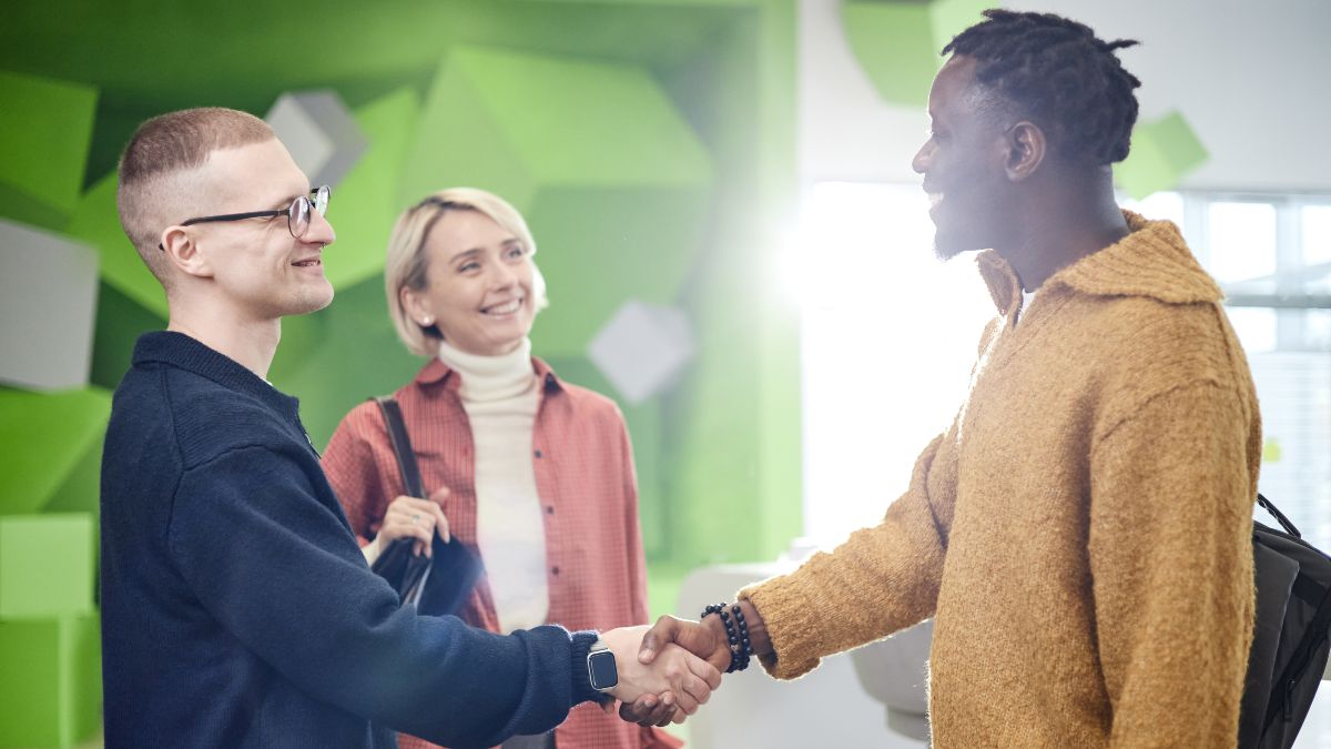 Zwei junge Männer im Büro schütteln sich die Hand und lächeln, während eine Frau im Hintergrund zuschaut.