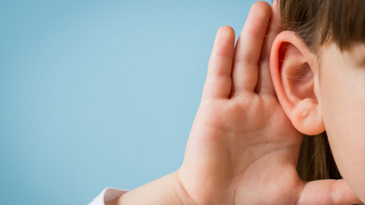 Vor blauem Hintergrund ist am rechten Bildrand in Nahaufnahme ein drittel des Gesichts eines Kindes zu sehen. Es hält sich eine Hand zum Lauschen hinter das Ohr.